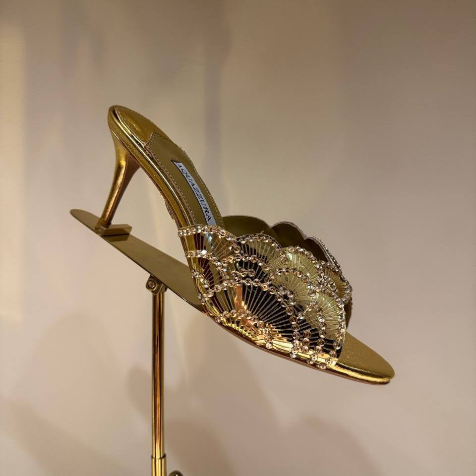 Gold high-heeled shoe with intricate design on a stand against a neutral background